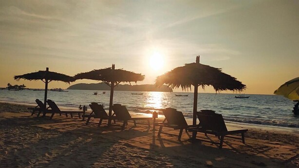 Une plage avec des chaises longues et des parasols en chaume, donnant sur une eau calme et un coucher de soleil.