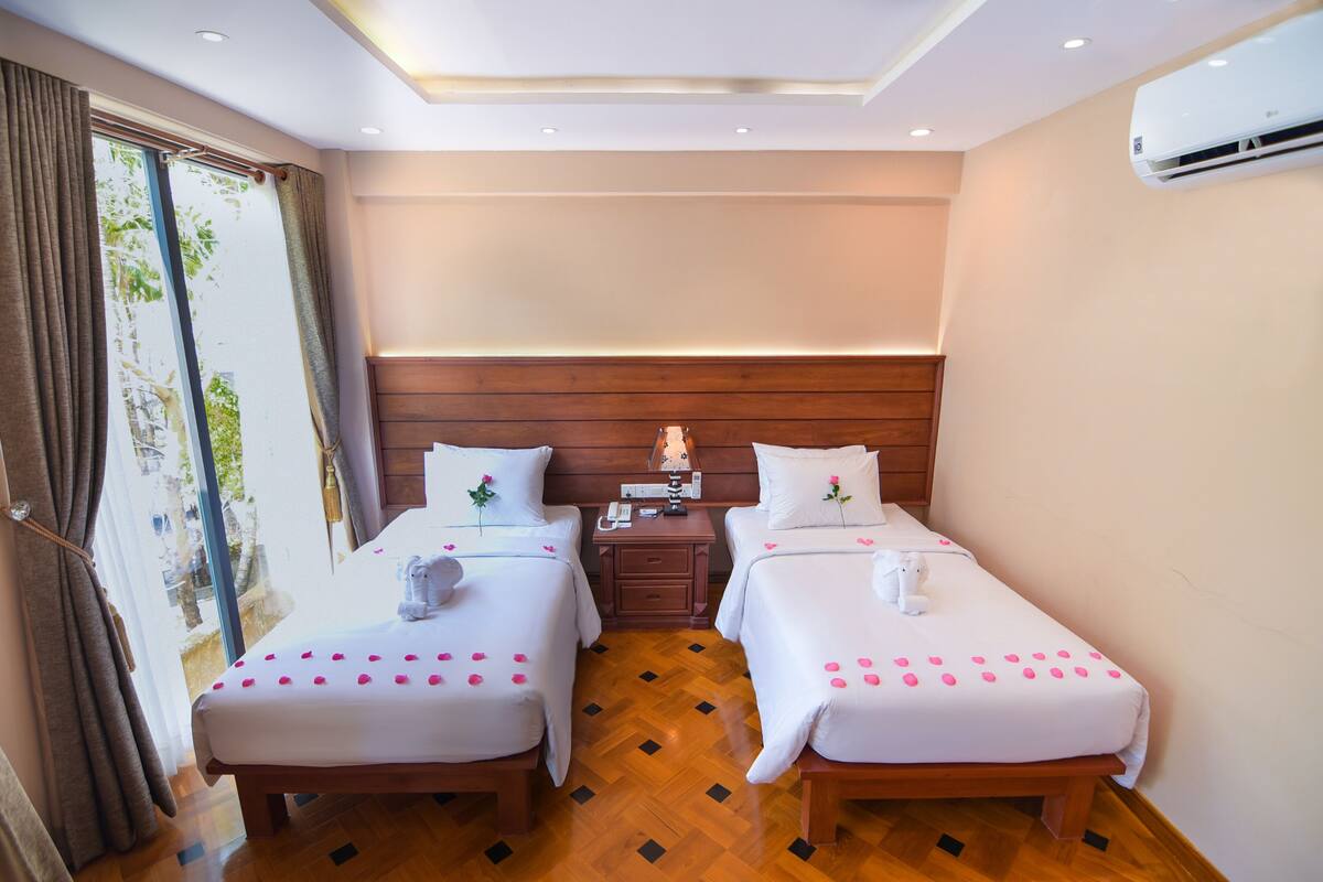 A hotel room with two beds, a wooden headboard, a nightstand with a lamp, and an air conditioning unit.