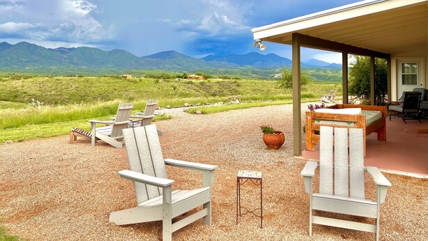 A covered outdoor seating area with Adirondack chairs and a table, overlooking a mountainous landscape.