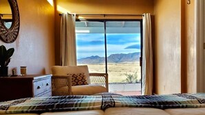 A bedroom with a large bed, a chair, a dresser, and a view of a desert landscape through a sliding door.
