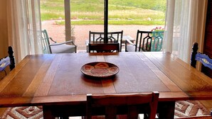 A wooden dining table with a woven placemat, surrounded by chairs, set against a backdrop of a mountainous landscape visible through large windows.