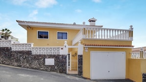 Un edificio amarillo con un balcón blanco, un garaje y un muro de piedra.