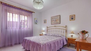 Un dormitorio con una cama, una araña, una mesita de noche, una lámpara y un florero con flores.