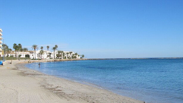 A beach with a clear blue sky, sandy shore, and white buildings with palm trees. - One bedroom apartment in Veneziola (La Manga del Mar Menor)
