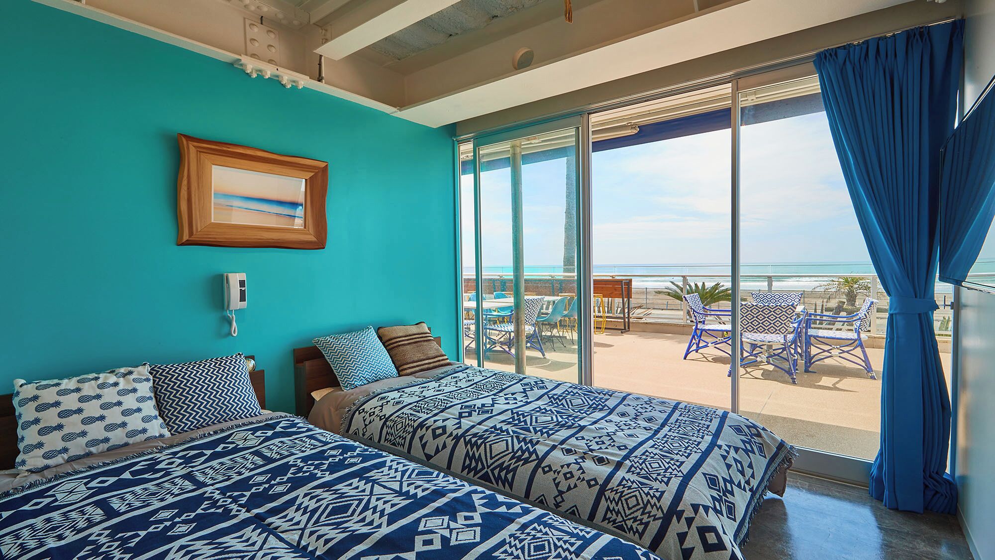 Ein Zimmer mit zwei Betten, einem Telefon, einem gerahmten Bild und Blick auf den Strand durch große Glasüren.