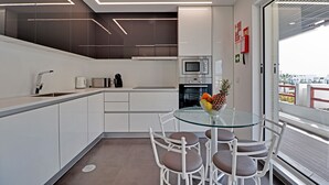 A modern kitchen with white cabinetry, a glass dining table, and a view through a large window. - MARINA LUXOR APARTMENT (Vilamoura)