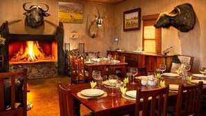 A restaurant with wooden tables set for dining, a fireplace with a roaring fire, and mounted animal heads on the walls. - Gannabos Guest House (Gariep)