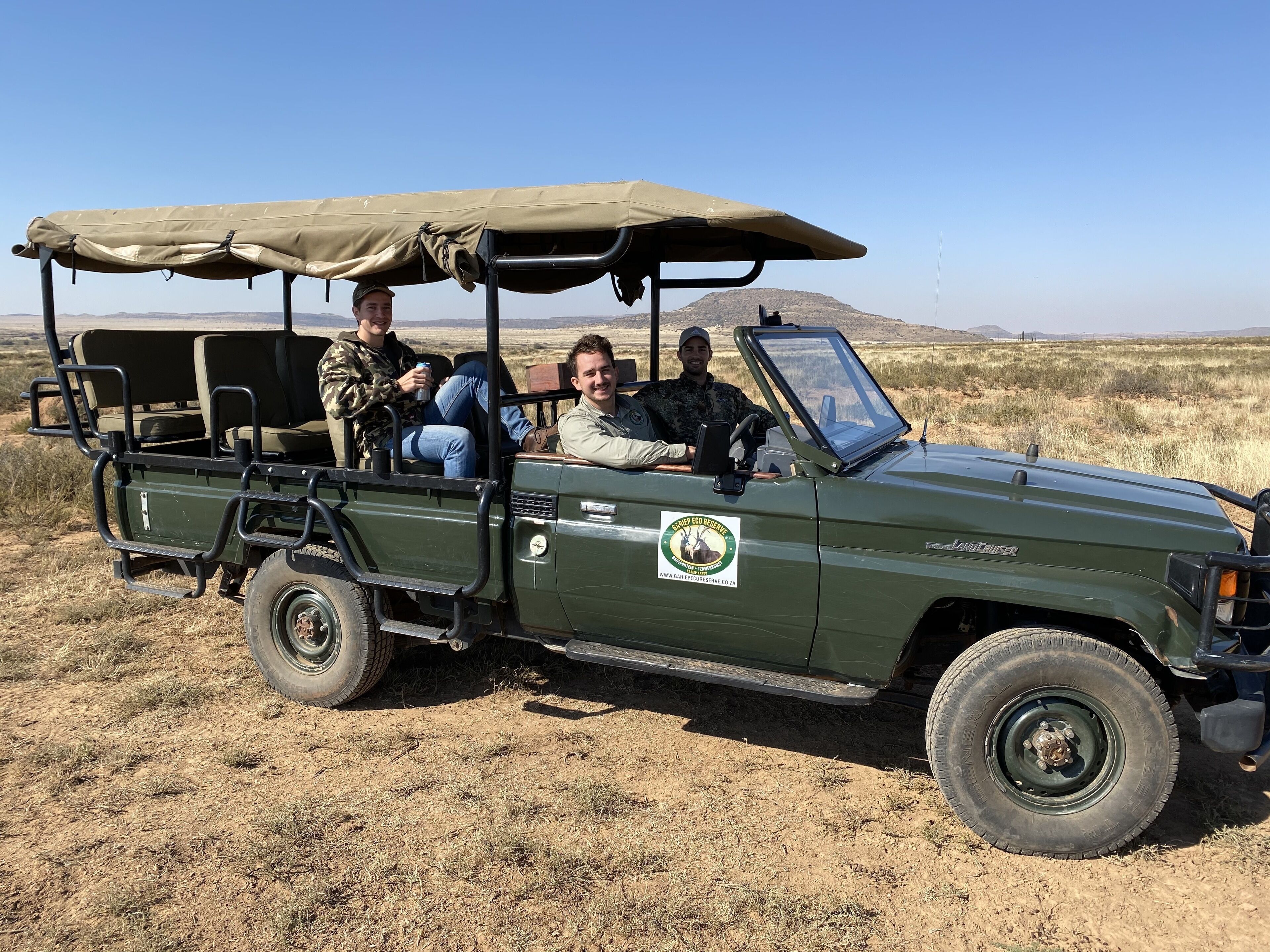 A green safari vehicle with a canopy, equipped with a side table and a steering wheel, parked in a dry grassland with mountains in the background.