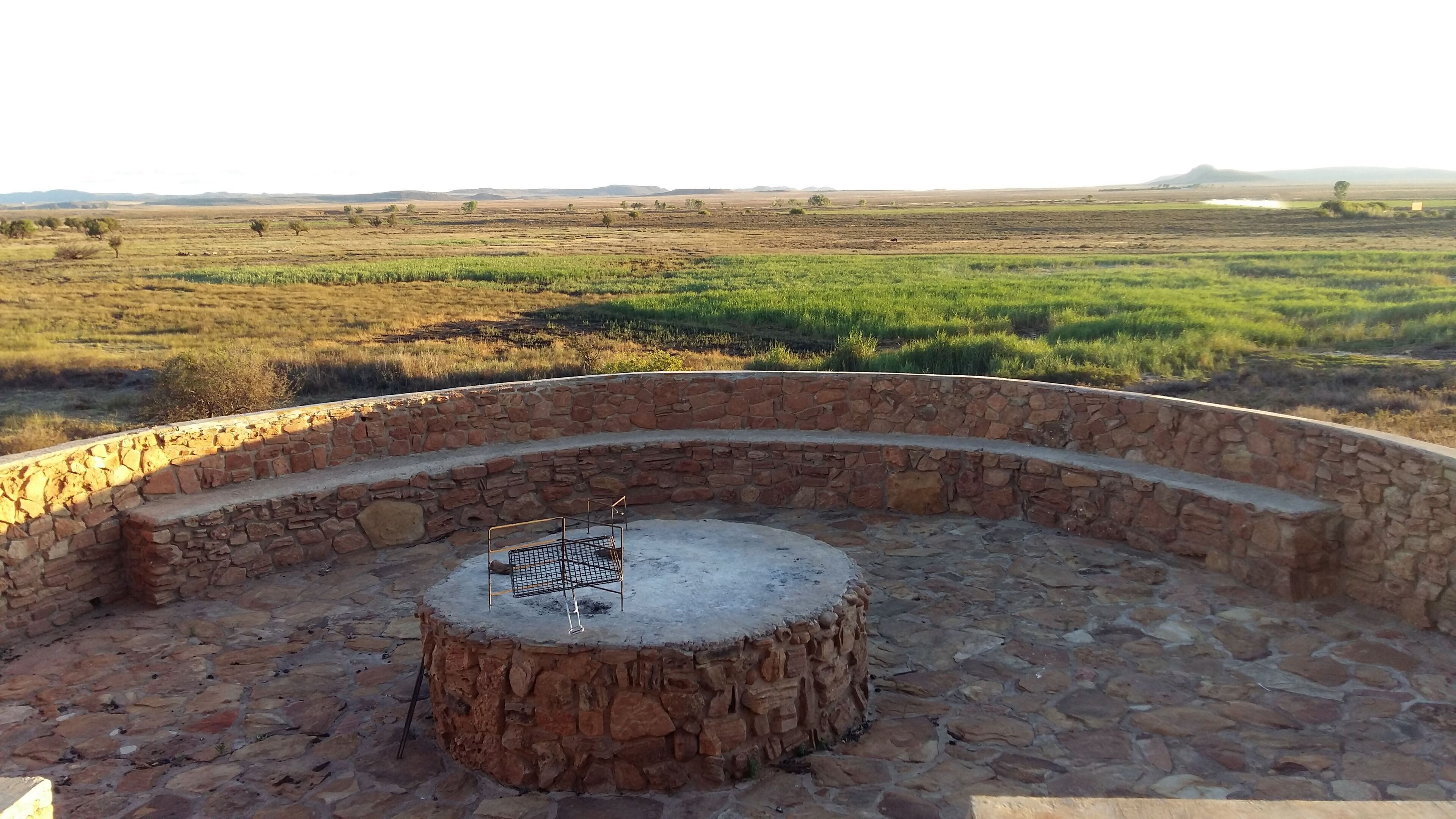 A stone-walled outdoor seating area with a metal grill table, surrounded by a natural landscape.