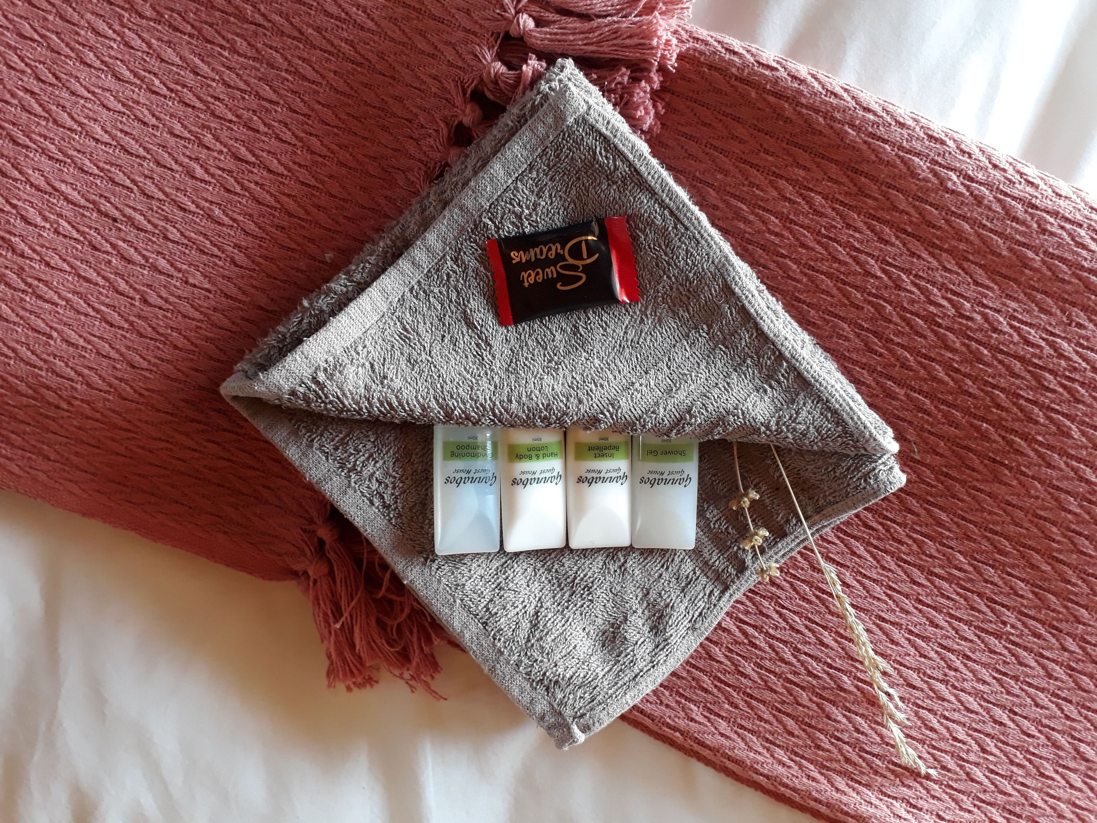 A grey towel with a black and red tag and three small white bottles on a bed with a red blanket.