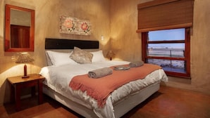 A bedroom with a bed, a nightstand, a lamp, a painting, a window with a view, and a wooden ceiling. - Gannabos Guest House (Gariep)