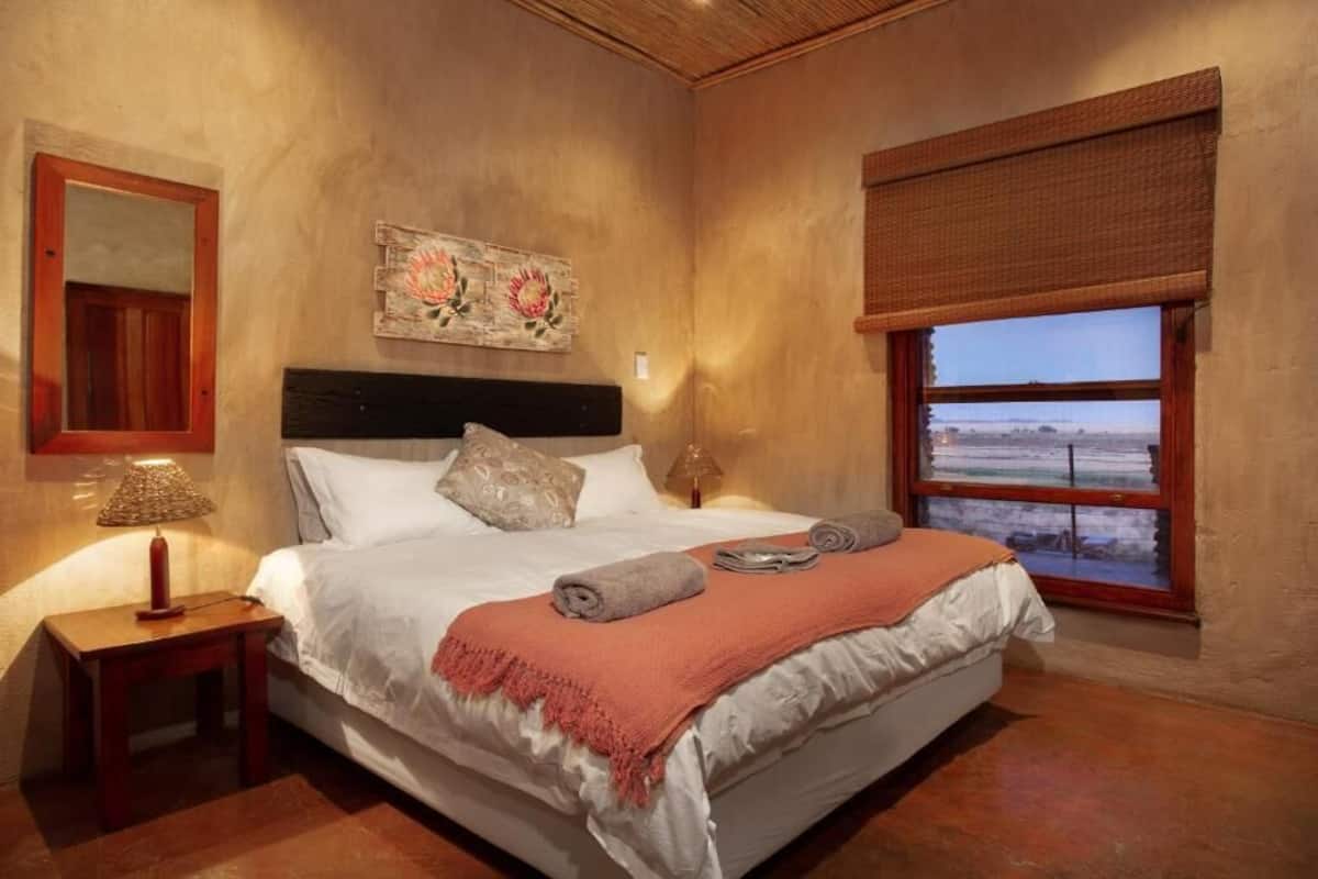 A bedroom with a bed, a nightstand, a lamp, a painting, a window with a view, and a wooden ceiling.