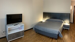 A modern hotel room with a flat-screen TV on a white stand, a bed with a grey headboard, and bedside lamps.