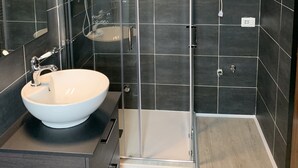 Moderni kylpyhuone, jossa on lasiseinällä varustettu suihku, valkoinen pesuallas ja tummat laattaseinät.