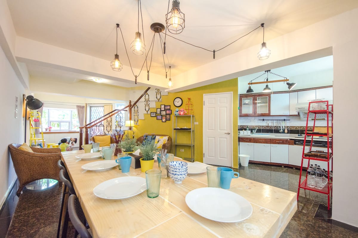 Un comedor con una mesa de madera dispuesta para cuatro, una cocina al fondo y una escalera que sube a otro nivel.