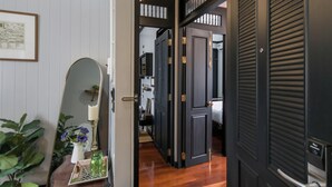 Un pasillo con puertas de madera oscura, una pared blanca con un cuadro enmarcado y un espejo que refleja un jarrón con flores moradas.