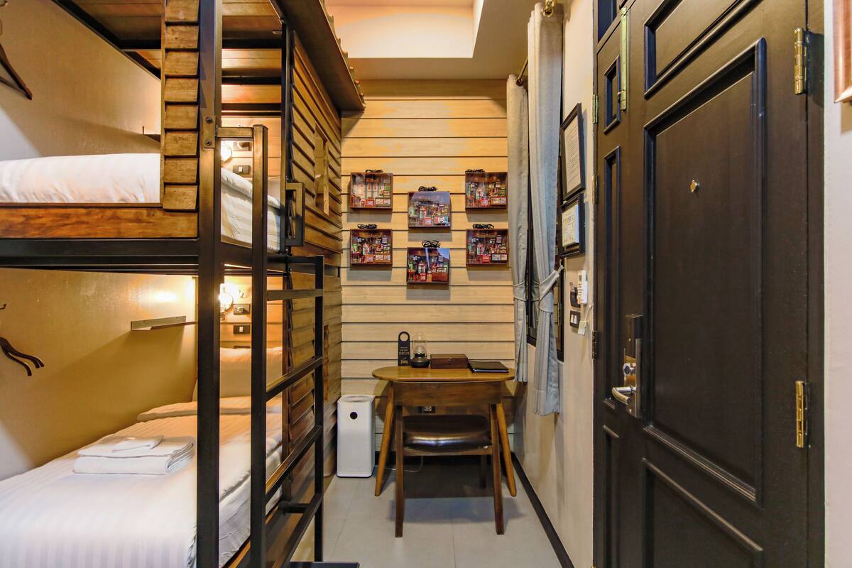 Un estrecho pasillo de hotel con literas a un lado y una pequeña mesa con una silla en el centro.