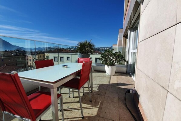 A rooftop terrace with a table and chairs, potted plants, and a clear view of the cityscape.