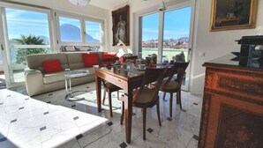 Un soggiorno moderno con tavolo da pranzo, divano e poltrone. Grandi finestre offrono vista sulla città e una montagna sullo sfondo.