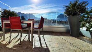 Terraço com mesa e cadeiras, uma grande planta em vaso e vista para montanhas.