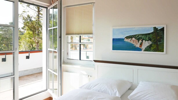 Une chambre avec une grande fenêtre, un tableau représentant un paysage côtier et un lit recouvert de draps blancs.