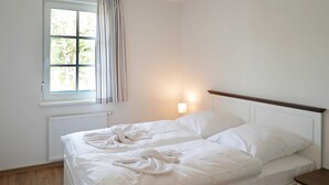 Ein sauberer, weißer Raum mit einem Einzelbett, einem Nachttisch, einem Fenster mit Vorhängen und einer Wandleuchte.