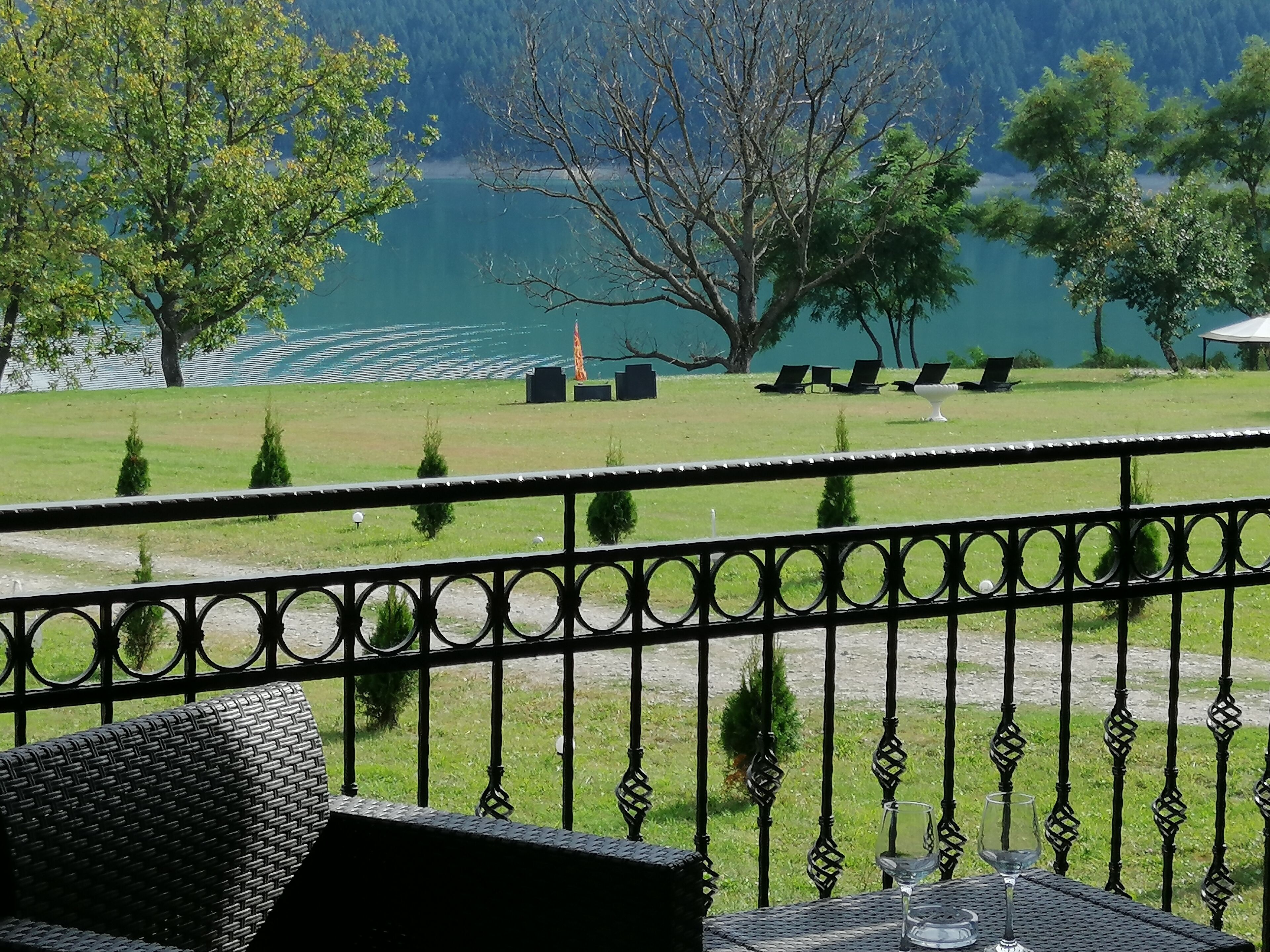 Un balcón con vistas a un lago, un área verde con árboles y mobiliario exterior.