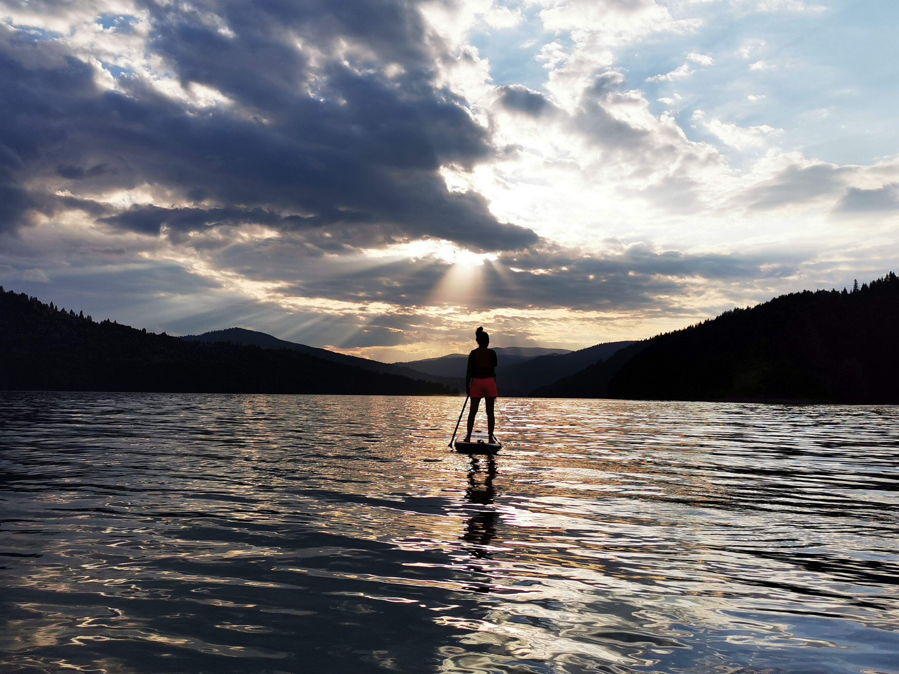 Una persona practicando paddleboard en un lago tranquilo con montañas al fondo y un cielo nublado.