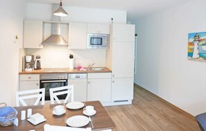 Une cuisine compacte avec des meubles blancs, un four à micro-ondes, une machine à café et un évier. On y trouve une table à manger avec des chaises et, au mur, un tableau représentant un phare.