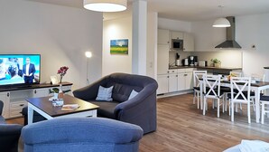 Un salon moderne avec un canapé, une table basse et un téléviseur. On y trouve également un coin repas avec une table et des chaises. La cuisine est visible au fond, avec ses appareils électroménagers et ses meubles de rangement.