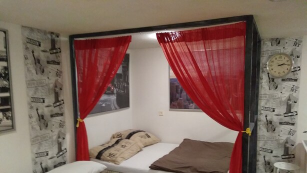 A small, single-bed room with a red canopy bed, a small table, and a clock on the wall. - Apartament w Centrum Noclegi Płock (Plock)