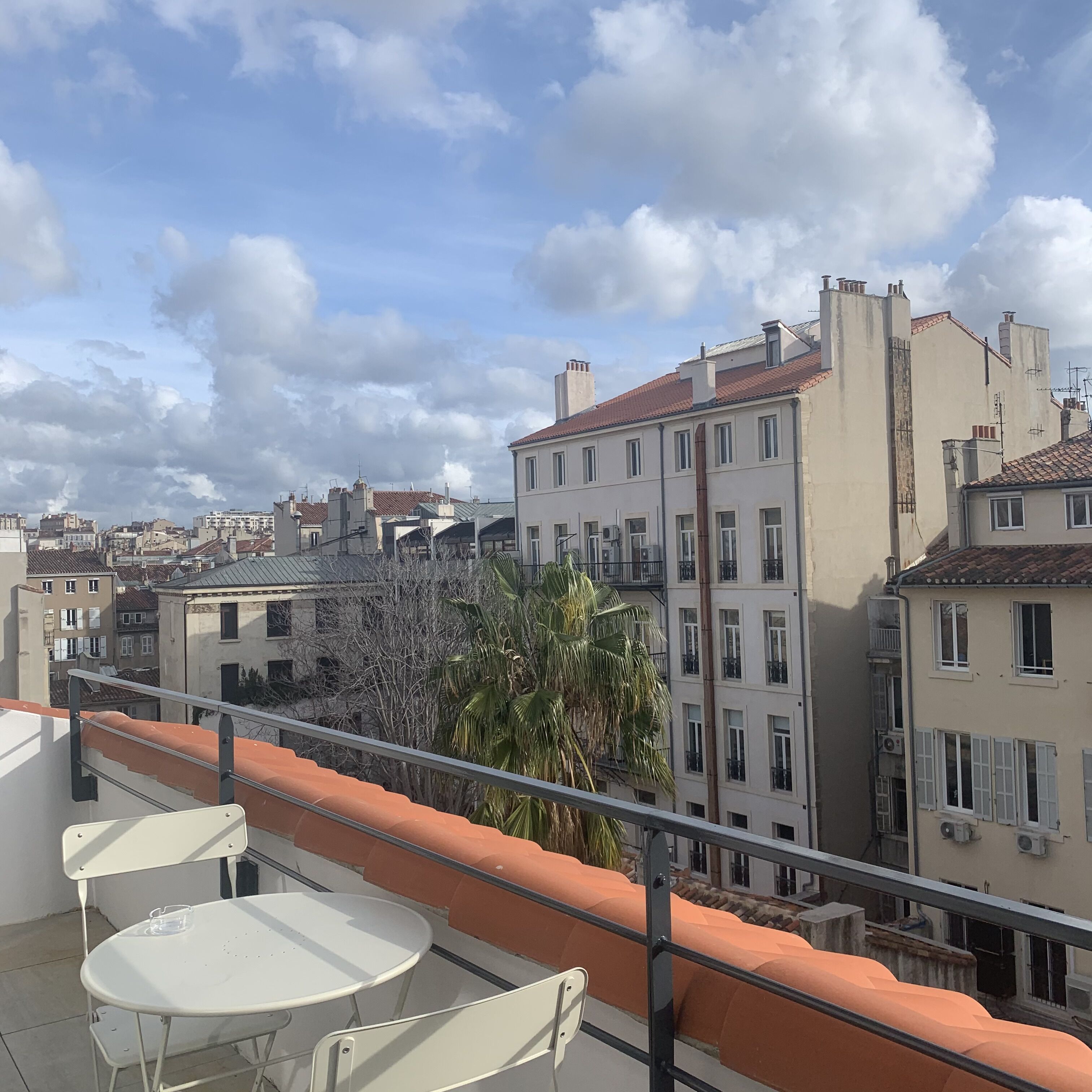 A terrace with a white table and chairs, overlooking a cityscape with buildings and a palm tree.