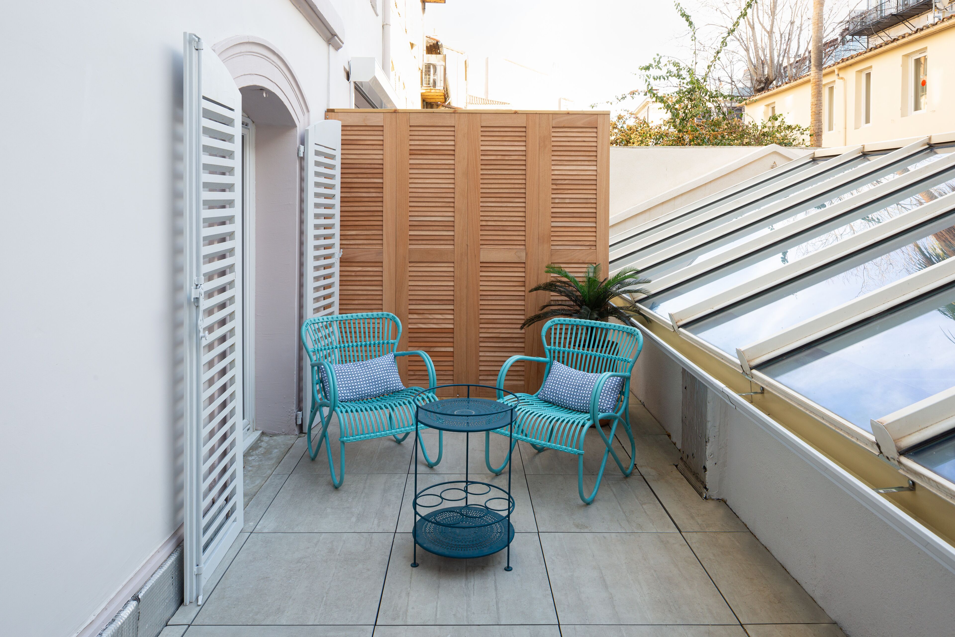 A small outdoor seating area with two turquoise chairs and a round table, a wooden partition, and a view of the sky through a skylight.