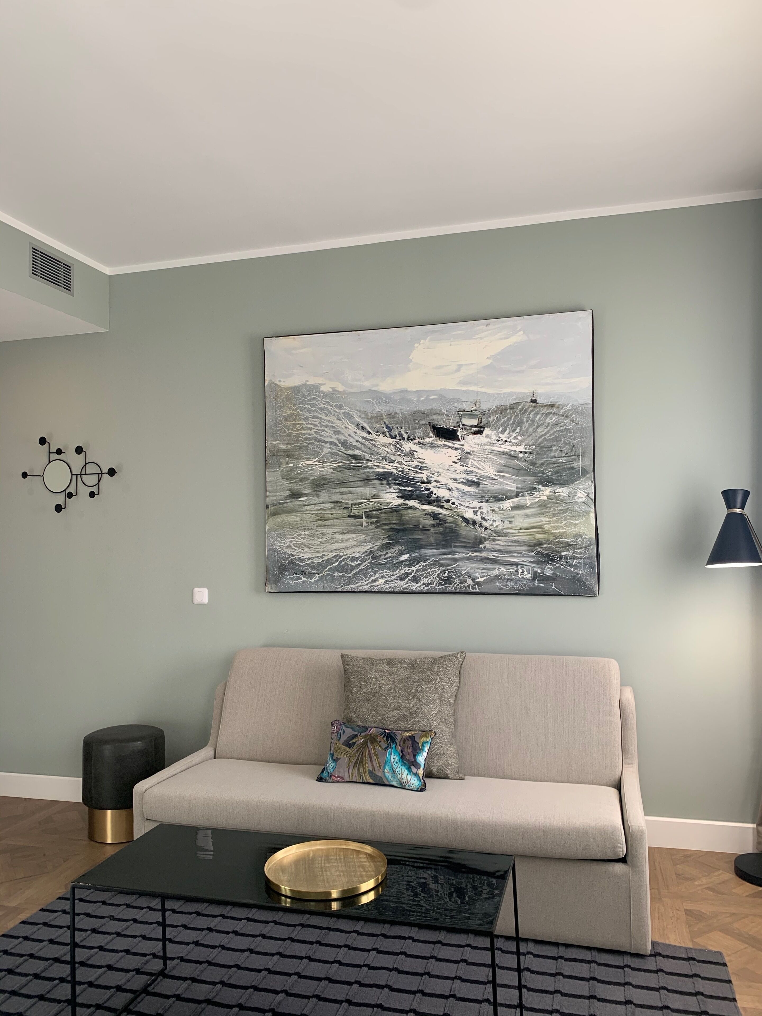 A living room with a beige sofa, a black coffee table with a gold tray, a patterned throw pillow, a wall-mounted abstract art piece, a floor lamp, and a small round stool.