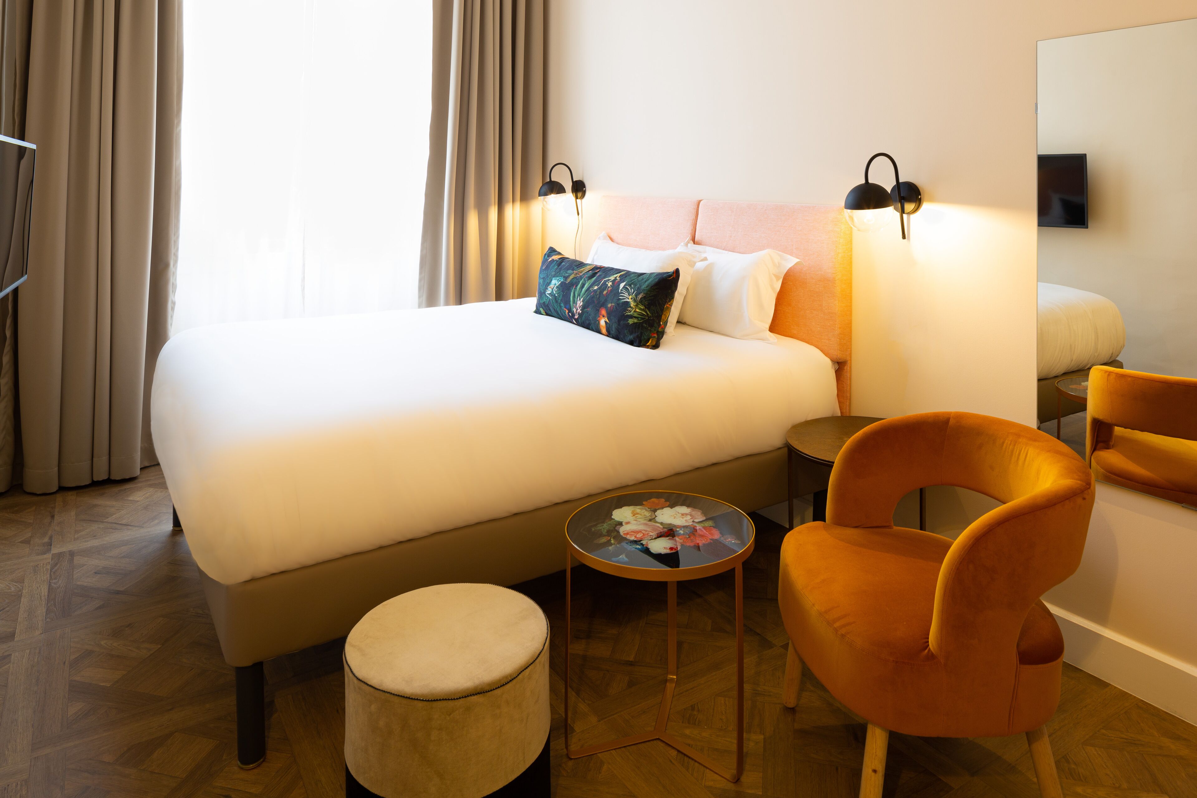 A hotel room with a large bed, a small round table with a flower arrangement, an orange chair, and a round ottoman.