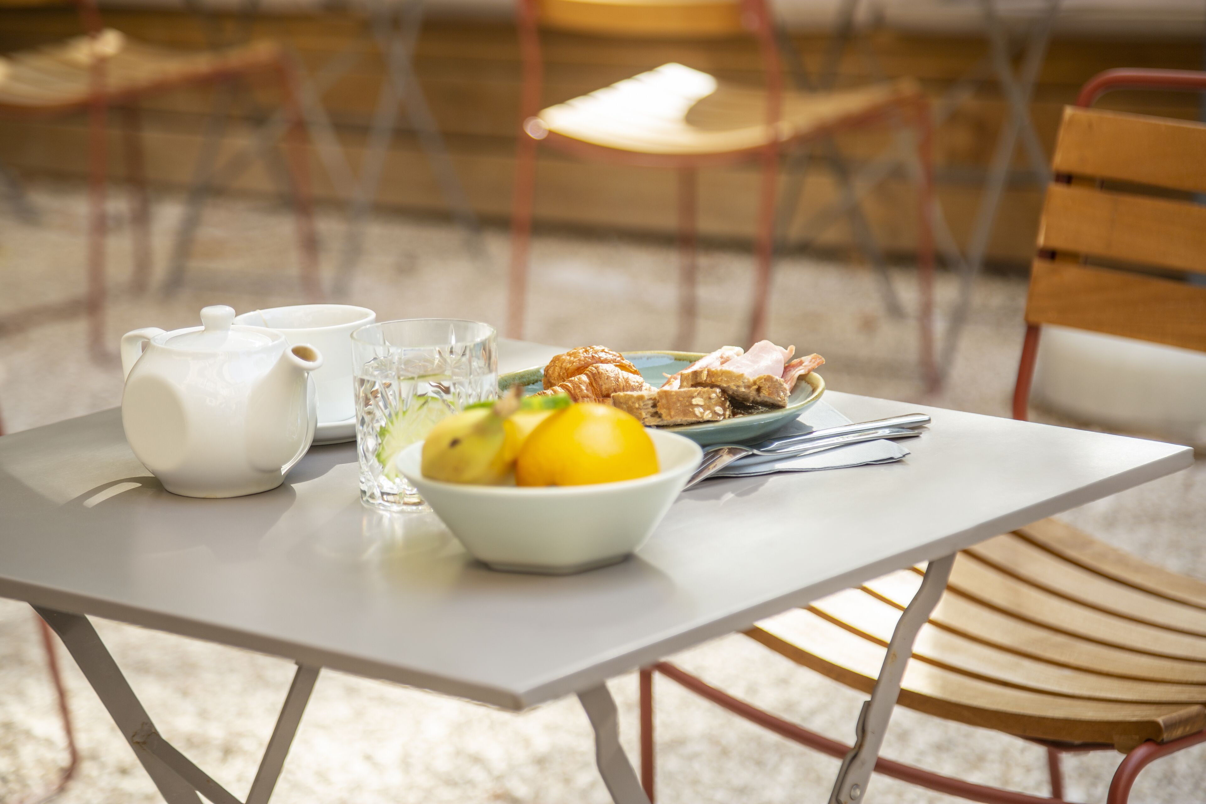 A table with a teapot, glass, bowl of fruit, and a plate with food.
