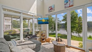A spacious living room with a large window overlooking a lake, a comfortable sofa, and a flat-screen TV mounted on the wall.