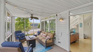 A covered porch with seating, a ceiling fan, string lights, and large windows overlooking a lake.