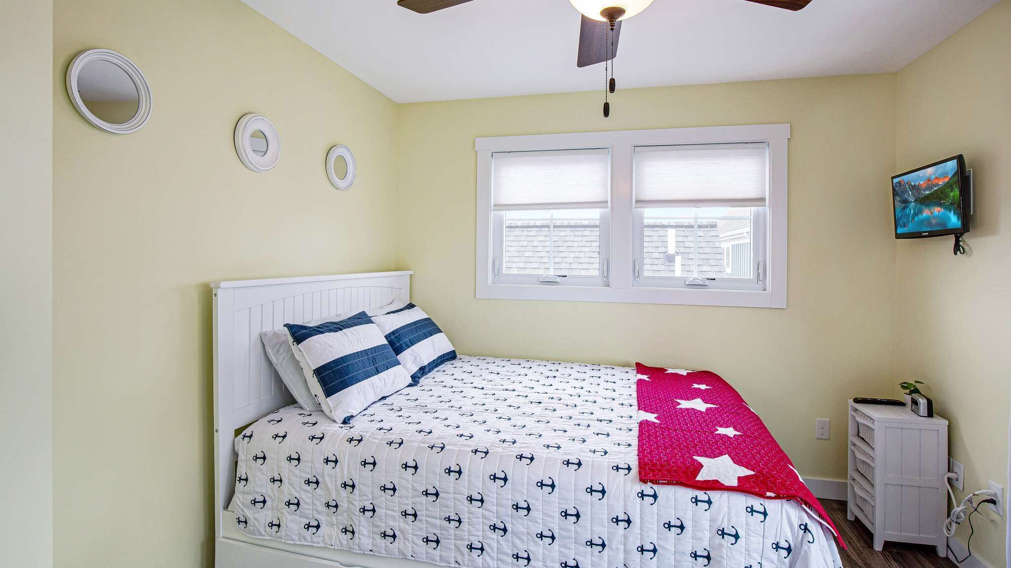 Une chambre à coucher avec un lit, un ventilateur de plafond, une fenêtre avec des stores, une télévision et une petite commode blanche.