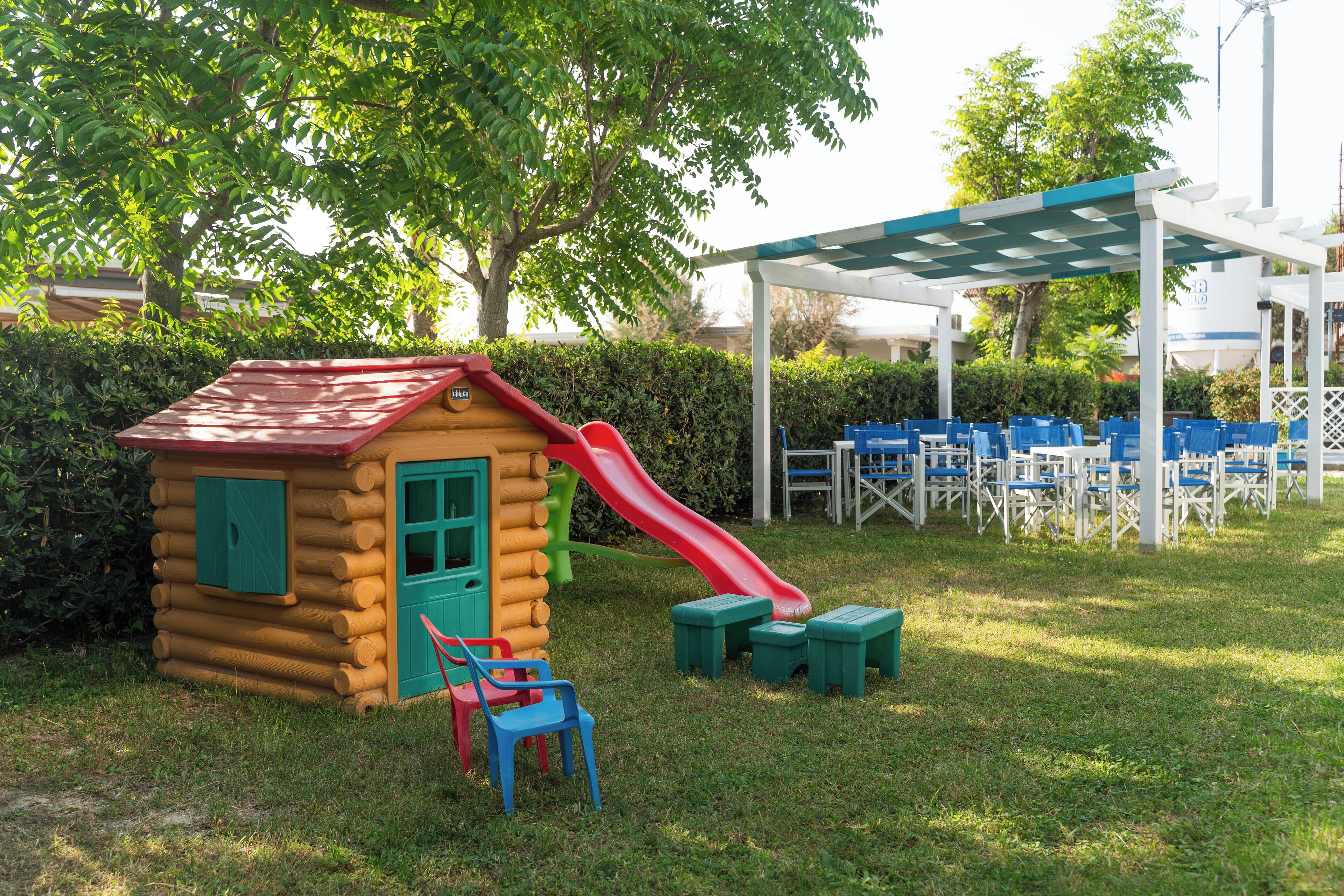 Una casita de madera tipo cabaña con un tobogán rojo, rodeada de arbustos verdes y una pérgola blanca con sillas azules.
