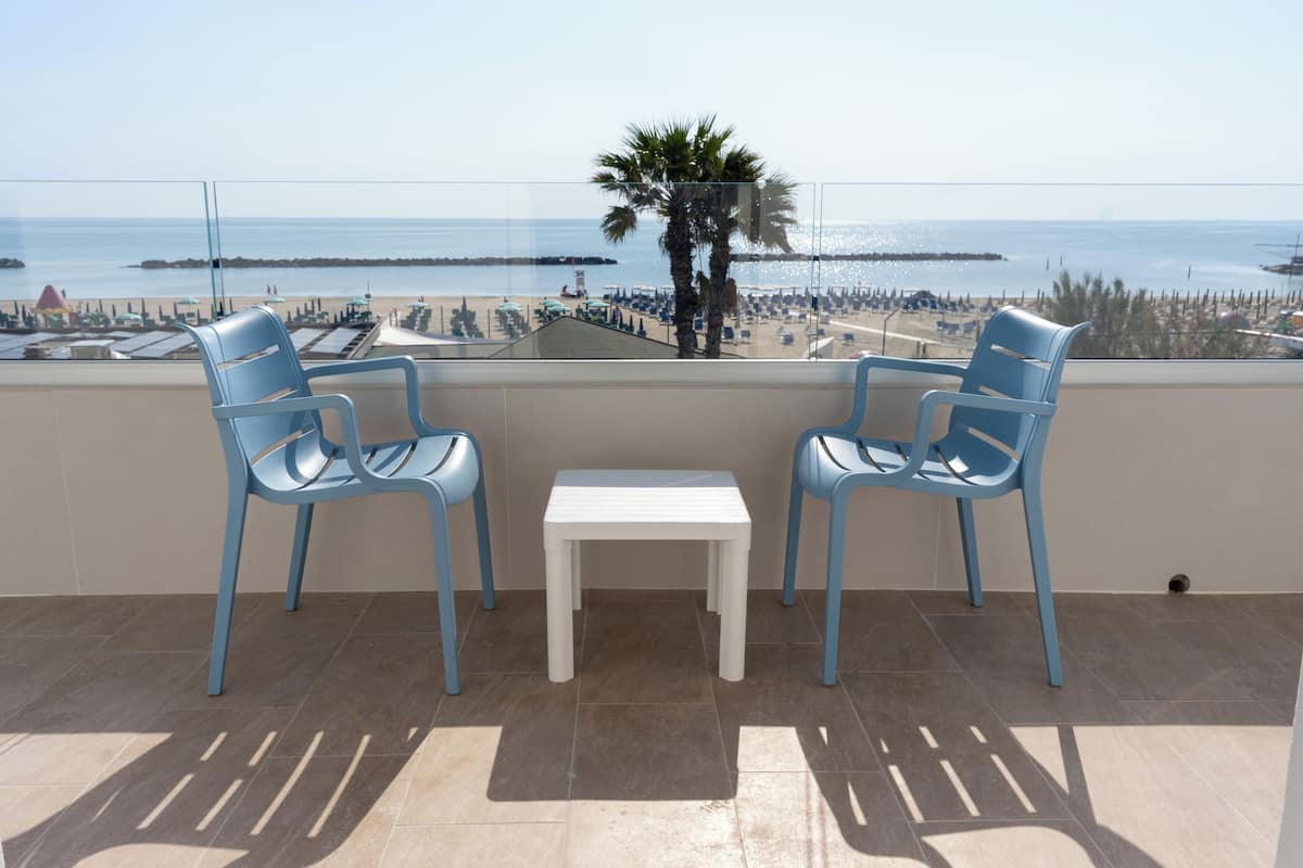 Un balcón con dos sillas azules y una pequeña mesa blanca, con vista a una playa que tiene una palmera y un muelle.