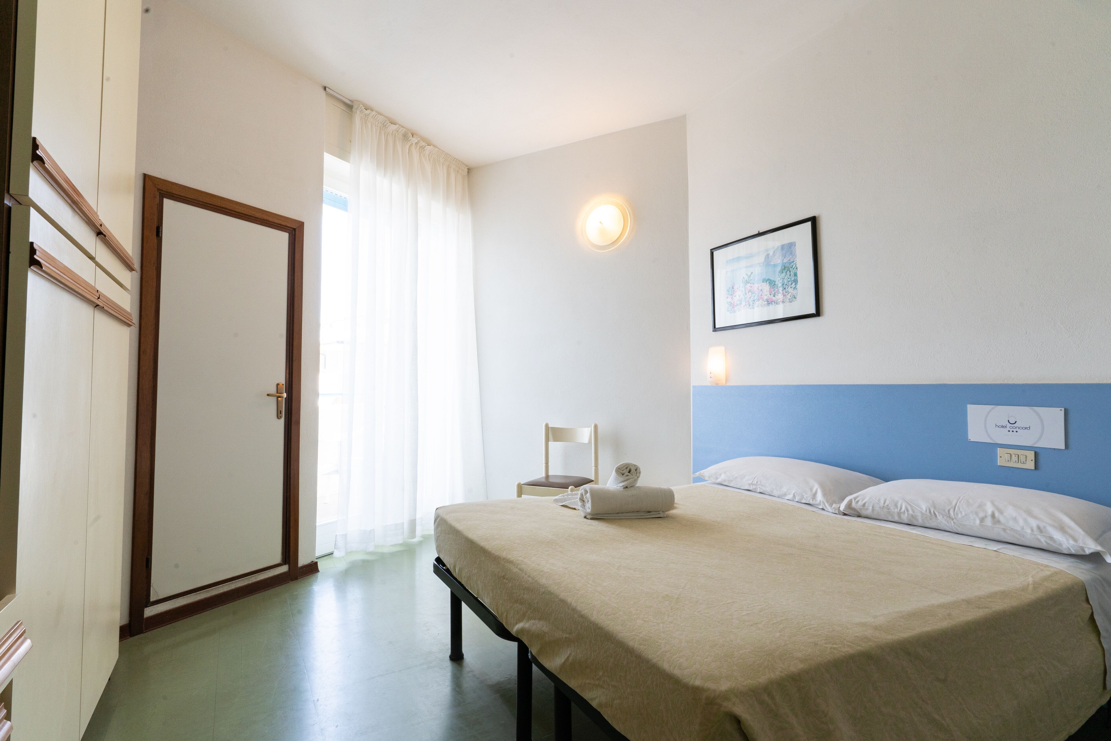 Ein Hotelzimmer mit Bett, Sessel, Wandlampe und einem fenster mit Vorhängen.