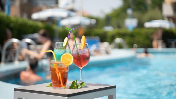 A poolside table with two drinks, a pool, and umbrellas in the background. - Hotel Concord (Ravenna)