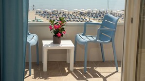 A balcony with two blue chairs, a small table with a flower vase, and a view of a beach with umbrellas and lounge chairs. - Hotel Concord (Ravenna)