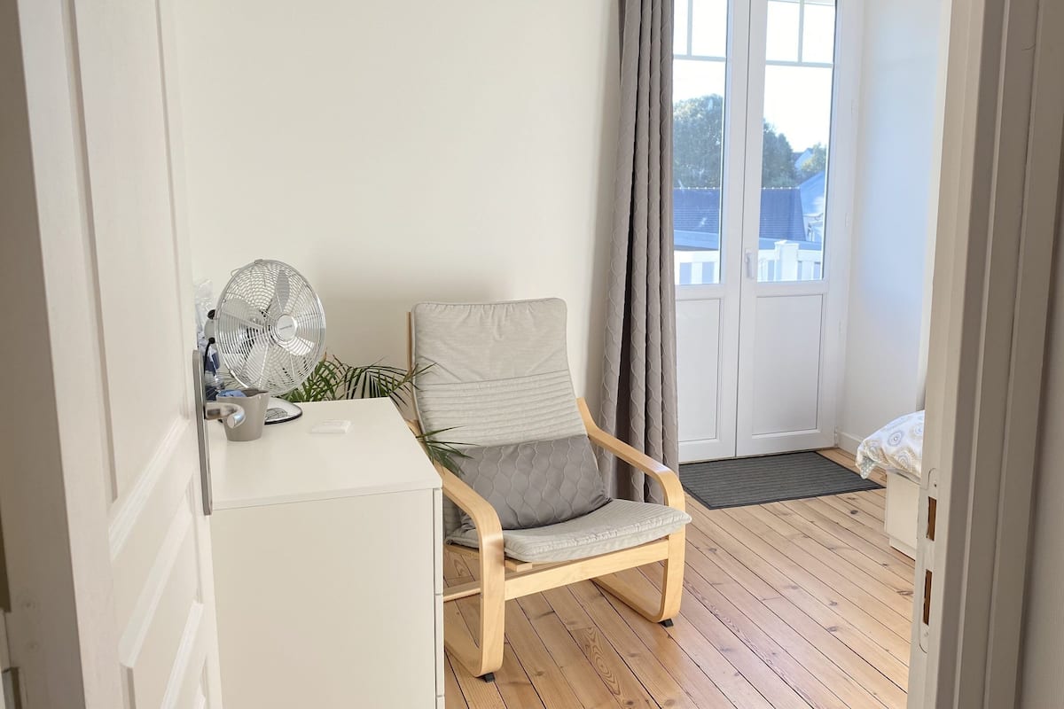 A room with a wooden floor, a grey cushioned chair, a white cabinet, and a window with curtains.