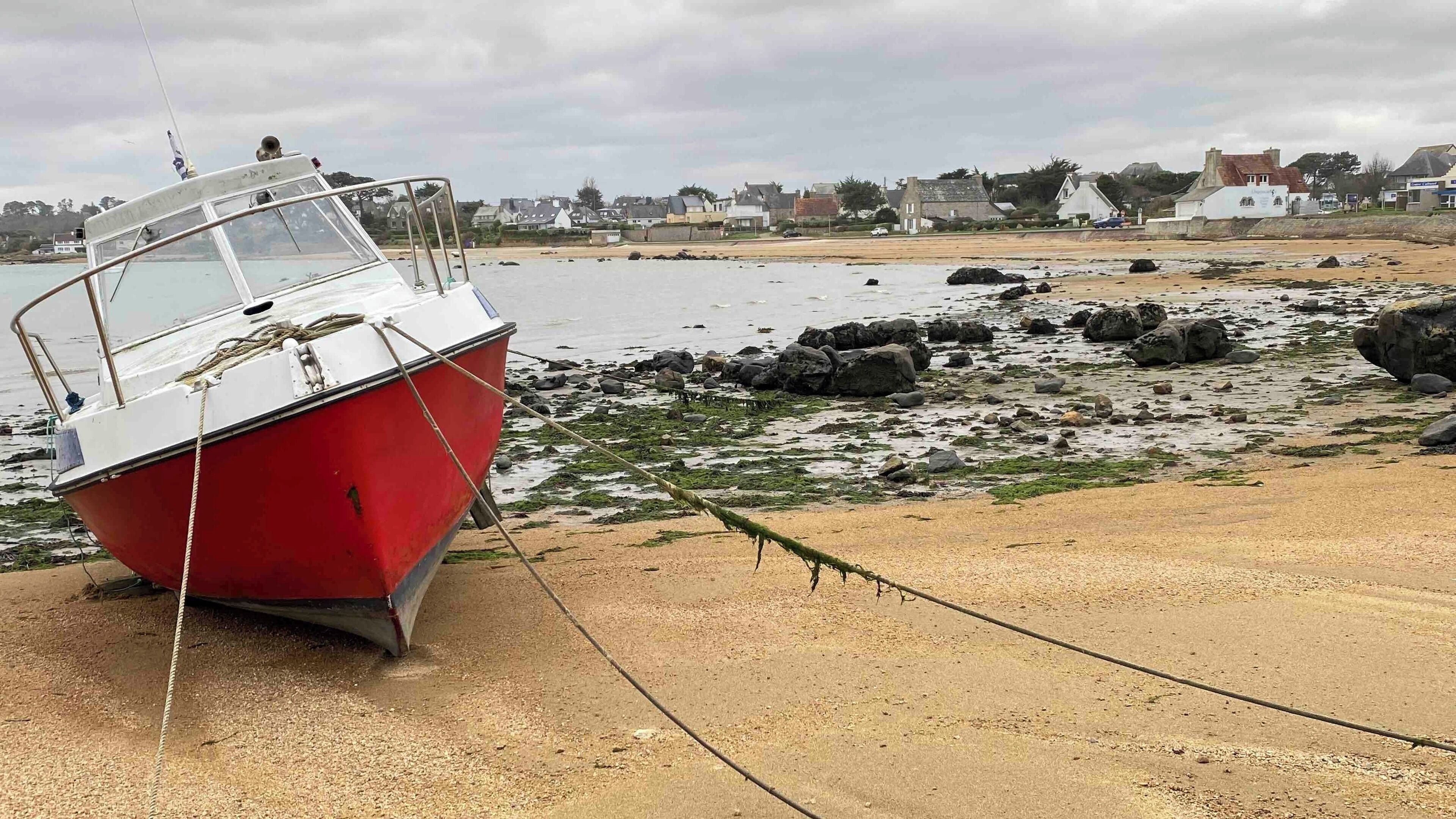 Un bateau rouge est amarré à un quai en béton, avec une c ôte rocheuse et des maisons en arrière-plan.