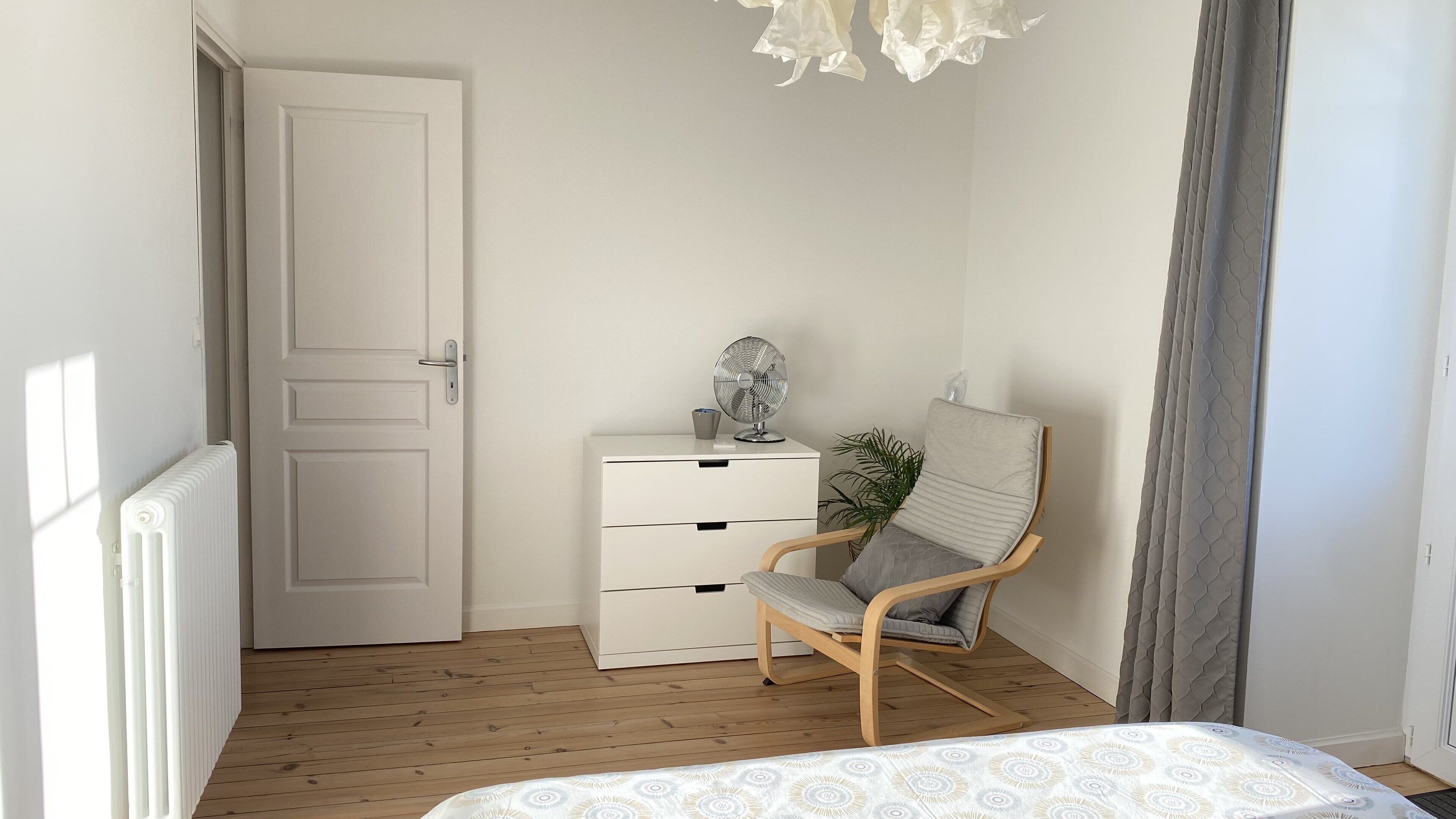 A bedroom with a wooden floor, a white dresser, a chair, a bed, and a radiator.