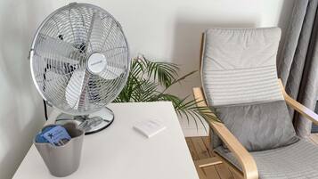 Una habitación con un escritorio blanco, una taza gris, un ventilador, una silla y una planta.