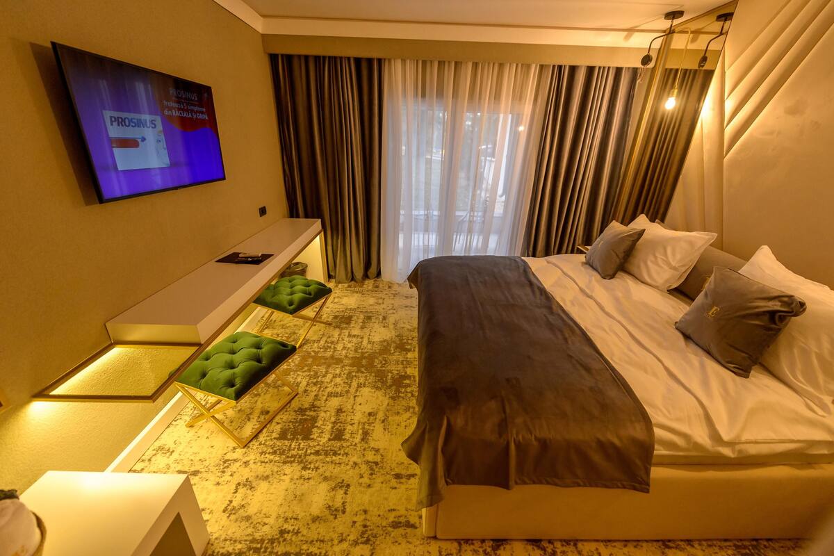 A hotel room with a bed, a desk, a TV, and a view of the outdoors through curtains.