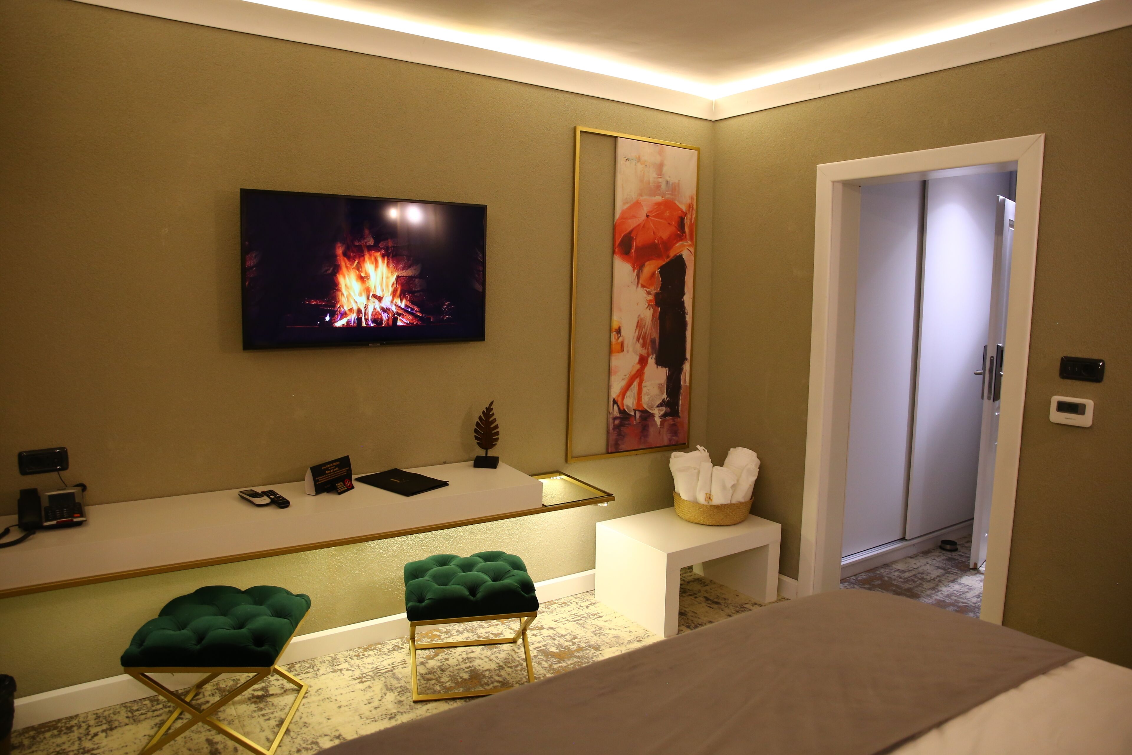 Habitación de hotel con televisión mostrando un fuego, un escritorio con teléfono y un libro, un otomano verde y un cuadro grande.